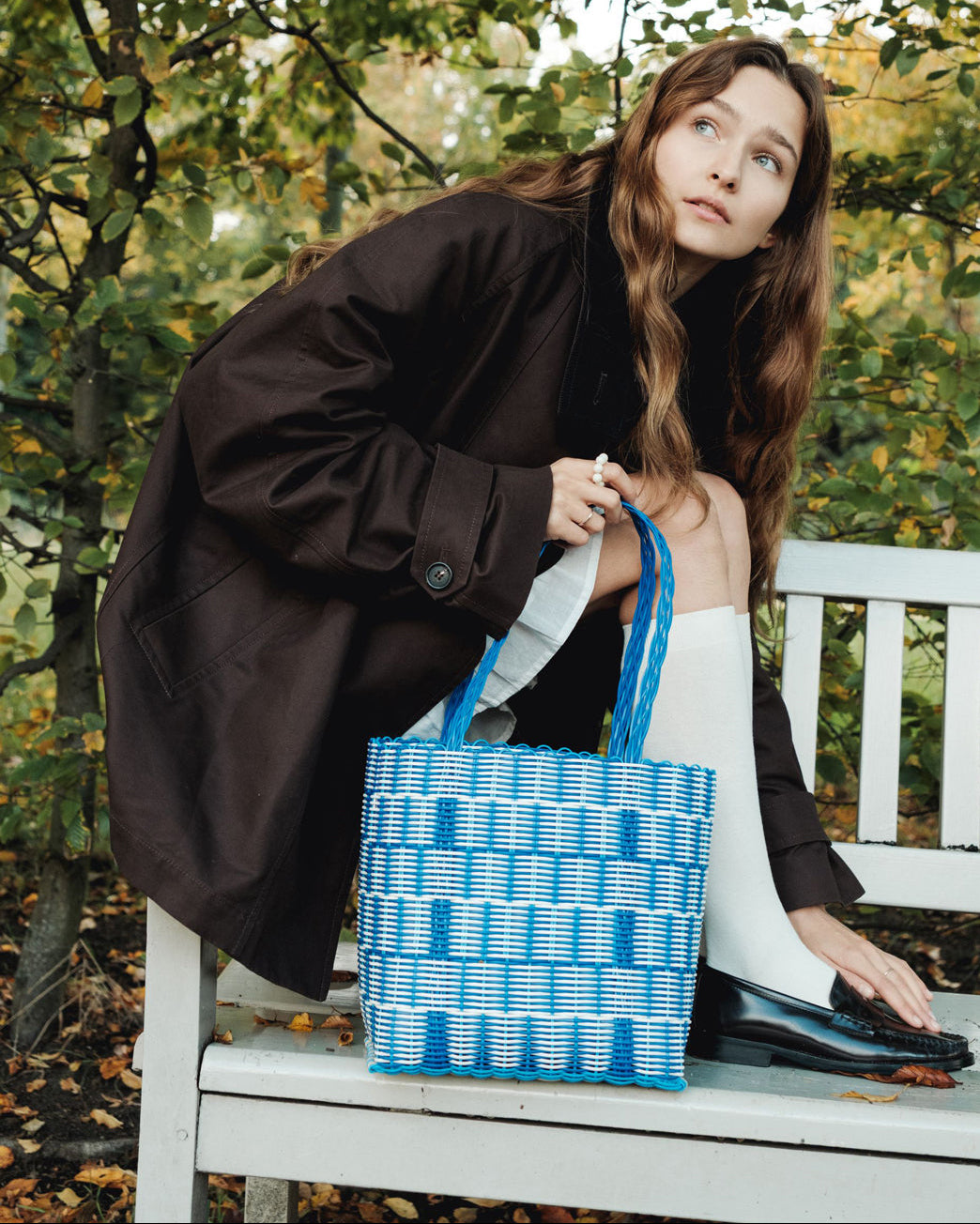Woman holding a blue patterned basket bag while sitting on a bench outdoors in a fall outfit.